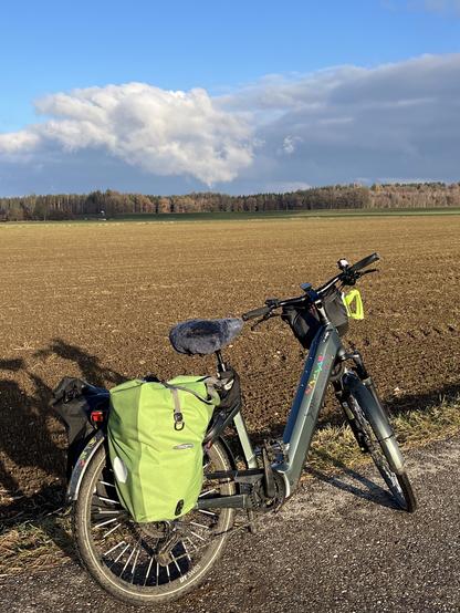 An electric bike parked beside a field, with a green pannier bag attached to the rear. There are clouds in the sky and trees lining the horizon.
Ein Pedelec, das neben einem Feld geparkt ist, mit einer grünen Tasche hinten. Es gibt Wolken am Himmel und Bäume am Horizont.