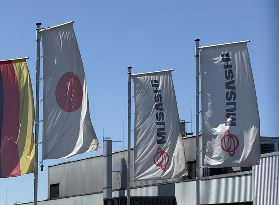 Drei Fahnen an Masten vor einem metallverkleideten Gebäude. Eine Fahne ist gelb-rot, eine weiß mit rotem Kreis, eine weiß mit dem Schriftzug "MUSASHI".