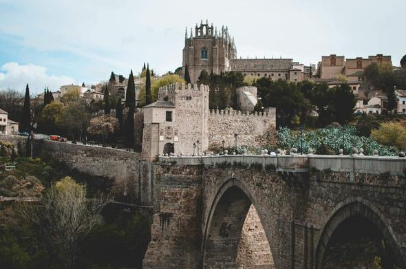 L'image montre un paysage urbain de la ville de Tolède, en Espagne. Un pont ancien en pierre, avec de grands arcs qui enjambent un ravin ou une vallée. Sur le pont, des lampadaires et des décorations blanches ajoutent une touche de charme à l'architecture.
À l'arrière-plan, une forteresse ou un château imposant se dresse sur une colline, surmontée d'une église à l'architecture gothique, avec de nombreux détails sur les fenêtres et les toits. La ville est construite sur une pente, et on peut apercevoir des maisons traditionnelles en pierre, ainsi que des arbres et des plantes qui ajoutent de la couleur et de la vie au paysage. Le ciel est partiellement nuageux, avec des nuages blancs flottants, créant une ambiance calme et douce.