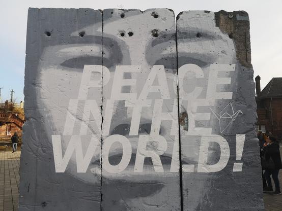 Das Graffiti zeigt in trauriges Gesicht in schwarz weiß hinter dem Schriftzug PEACE IN THE WORLD!
Der Untergrund sind drei Segmente der Berliner Mauer, die die Graffiti Künstler Gruppe 3steps aus Gießen erworben auf dem Gießener Bahnhofsvorplatz aufgestellt hat. Anlassbezogen wechseln die Botschaften von Zeit zu Zeit.