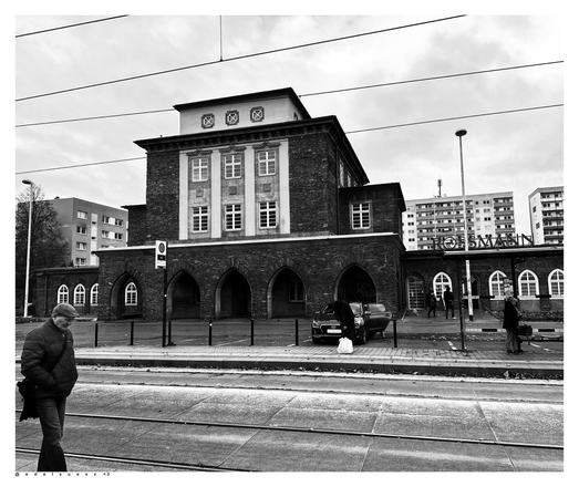 Aufnahme in schwarzweiß, das Empfangsgebäude des Flughafen, in Betrieb gegangen in den 1920er Jahre, genutzt bis Ende der 1960er Jahre, davor Gleise der Straßenbahn und ein ins Bild laufender alter Mann mit Schirmmütze, dahinter eckige Wohnblöcke aus DDR Zeiten