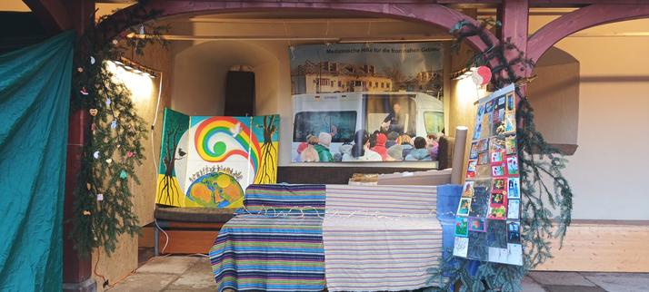 Ein fast fertiger Weihnachtsmarkt-Stand in Arkaden an einem alten Gebäude. Links etwas Tannenreisig mit Christbaumschmuck, hinten das Friedens-Flügelbild, rechts daneben ein riesiges Plakat im Querformat, auf dem der Arzt Dr. Matthias Werner bei einem Einsatz in der Ukraine gezeigt wird. Vorne Thekentische mit bunten Tüchern. Rechts Tannenreisig und eine Übersicht an Postkarten.