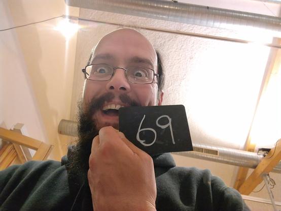 crazy looking bearded guy with balding head and glasses holding a table number...it is the number 69 (sixty nine)