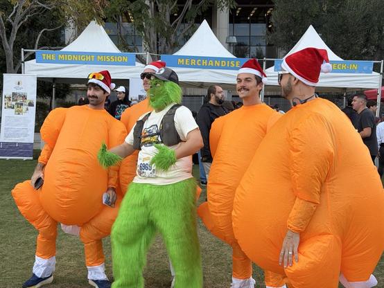 4 men in Santa hats and inflatable orange turkey costumes, standing with a person in a Grinch costume, who is wearing a Turkey Trot t-shirt over the green fur.