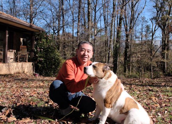 A man in an orange sweatshirt and blue jeans squatting in the yard at home holding his white and brown dog. Blue sky through trees on the background, a house on the left.