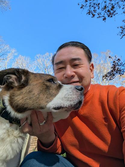 A phone selfie of a man in an orange sweatshirt forcing the big head of a white and brown dog into the frame. Blue sky and tree branches in the background