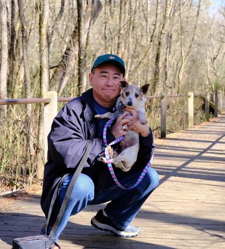 A man with a green cap squatting and holding his white and brown Jack Russell Terrier on a board walk in a park. 📍Heritage Park, GA