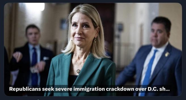 Rep Miller stands in a hallway, looking directly at the camera with a slight smile. Two men are partially visible in the background, one gesturing and the other appearing attentive. The setting suggests a formal or political environment.