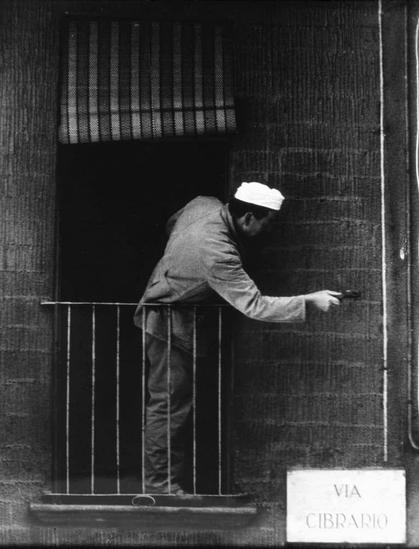 The image is a black and white photograph showing an individual leaning out of a window, reaching towards the door handle. The person appears to be wearing a suit with a light-colored hat or head covering, suggesting they could be in some form of professional attire possibly linked to the food industry due to this style being reminiscent of what one might wear while working as a baker at that time period.

The individual is interacting with something outside through an open window which has metal bars across it. The background includes part of a building wall and a sign on the exterior, indicating "Via Cibraio," suggesting that this location could be in or around a neighborhood known for its connection to food production or distribution.

This photograph captures what seems like a moment from daily life with an emphasis on labor rather than leisure activity. The image is noted as being part of Bonis' collection and associated with the Ferrania brand, likely indicating it's related to industrial film photography used during that era in Italy, particularly around 1964.

The black-and-white palette of this photograph suggests its age or a deliberate stylistic choice by the photographer. The contrast is high, providing sharp details on textures like the building material and clothing fibers.