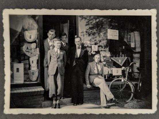 A vintage photograph with a white border and decorative, scalloped edges. In the photograph, a group of five men dressed formally is standing in front of a storefront with large glass display windows. Several signs are visible in the window. One prominent sign reads, "F.A. STERK & Zn." 

This shop belonged to my great-grandfather and grandfather. The windows are packed with merchandise, including textiles, linens, and household goods. My father stands partially in the shadows of the doorway behind his father and brothers.