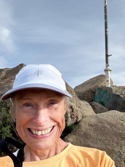 A woman in a yellow shirt and white baseball cap is standing on top of a mountain. Behind her are huge boulders and a metal pole to mark the top. Right after Thanksgiving, they used this pole to erect a Christmas tree made of strings of lights. Cerro San Luis in San Luis Obispo, California