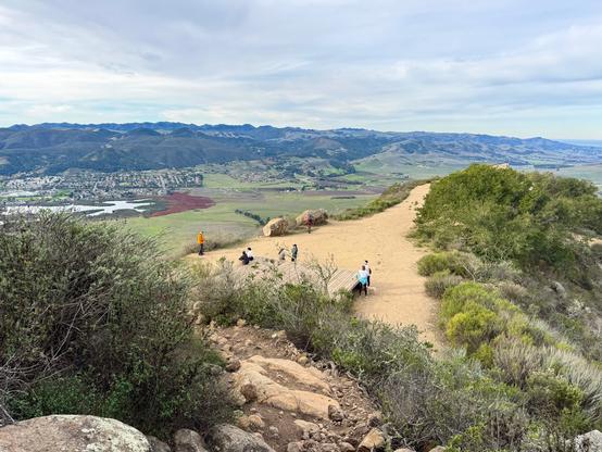 Looking down at the trail that leads to the top of Cerro San Luis in San Luis Obispo, California. The trail goes across the ridge at the top, and there is a wooden deck at the top as well. There are several people gathered around the deck as they rest before going back down. The mountain is mostly shrubs with some trees. The view in the distance shows a valley with a lake (Laguna Lake) and mountains beyond. The sky has high, thin clouds, so the sunlight is filtered.