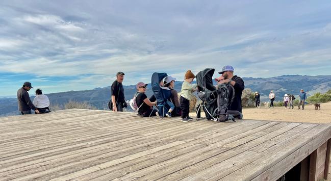 Groups of hikers are resting at the wooden deck at the top of Cerro San Luis in San Luis Obispo California. More hikers are approaching from the trail leading up to the top. There are all ages of people, and a dog. The sky is thin clouds.