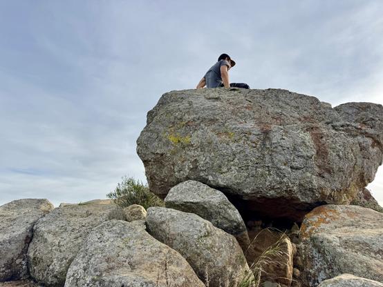 A man sits on top of one of the huge boulders on top of Cerro San Luis in San Luis Obispo, California. He has a baseball cap on and is looking down at the view below. The sky is thin clouds.
