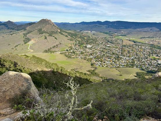 A view from the top of Cerro San Luis in San Luis Obispo, California. In the foreground are boulders and brush. Beyond the mountain is another peak, Bishop Peak, and the city of San Luis Obispo at its base. There are more mountains in the distance.