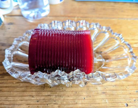 A cylindrical blob of jellied cranberry still in the shape of the can sits in the middle of a crystal dish. The dish sits on a sunny wooden table.