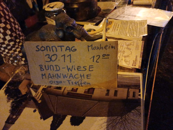 Auf einem Infotisch der Mahnwache in Köln-Kalk steht neben Broschüren und Firguren ein Pappschild: "Sonntag 30.11., 12 Uhr, Manheim, BUND-Wiese MAhnwache Orga-Treffen"