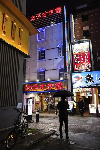 A quiet street bathed in the red and purple neon lights of a karaoke place in Shimbashi, in Tokyo, at night. In the foreground the silhouette of a man in a dark costume standing and holding a black umbrella. Opposite him next to the Karaoke, a restaurant waiter, also dressed in black, is taking a break in front of his shop.