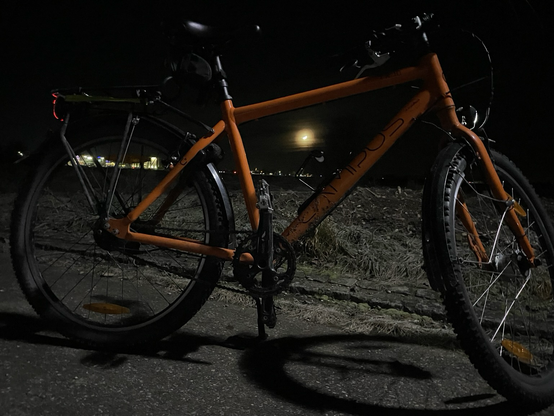 Ein Fahrrad steht an einer Wiese. Es ist Nacht, durch das Rahmendreieck scheint der tiefstehende Halbmond.