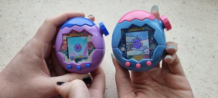 close up of scremlin and I's hands holding our tamagotchis. Scremlin has a purple and blue tamagotchi paradise and I have the same in blue and pink. Scremlin has a purple bumblebee like tama in the sky and I have a have purple beaver like tama in the ocean. The camera is focused on my tama who has an excited expression. Scremlin's is also excited, but a little blurry.