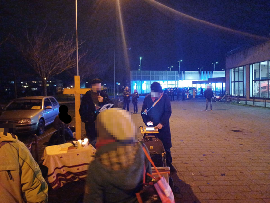 An einem improvisierten Altar auf einem Parkplatz mit Kerzen und einem gelben Kreuz stehen zwei Männer, davor weitere Personen. Im Hintergrund stehen Polizist*innen vor einem Parkhaus im Dunkeln
