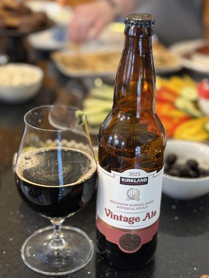 A snifter glass half full of dark beer. The brown bottle has a white label with a dark red lower band. A plate of vegetables is in the background