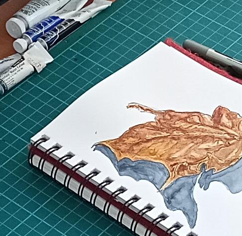 A cropped photo of a green cutting mat that has a spiral bound sketchbook on it.

The sketch is open at a page with an ink and watercolour painting of a leaf.