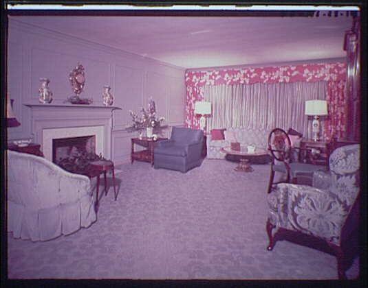 The image depicts a well-appointed sitting room within a hotel suite, circa the early to mid-20th century. The space features soft pink and cream hues on the walls, complemented by rich red floral patterns in curtains that frame one wall of large windows dressed with sheer white drapes allowing natural light into the room.

Two armchairs upholstered with intricate fabric designs occupy opposite sides of a carpeted floor, inviting conversation between guests. A plush sofa is situated against another side, near an ornate fireplace mantel decorated with several decorative items and flanked by two standing lamps adorned with red shades that echo the curtains' floral motif.

In front of the seating area stands a wooden coffee table holding various small objects including what appears to be books or magazines, alongside possibly some personal belongings. The room is furnished further with end tables on either side of each armchair, featuring items such as vases and perhaps flower arrangements adding to the elegance of this setting.

The space exudes comfort and classic luxury through its tasteful decor choices, reflecting a bygone era's design sensibilities.