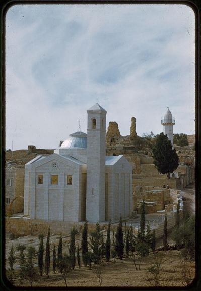 The image depicts a serene and picturesque scene of an ancient architectural complex, possibly from the Middle Eastern region. It features a prominent white stone church with distinctive religious iconography, including crosses on its facade and windows adorned with golden embellishments that suggest Christian influence.

Surrounding this central structure are remnants of historical buildings and fortifications in various states of ruin or preservation. To the right stands an imposing dome-covered tower, possibly part of another mosque or a minaret, indicating Islamic presence nearby. The landscape is dotted with sparse trees and shrubs, enhancing the arid ambiance typical of desert regions.

The overall composition conveys a sense of historical continuity amidst natural beauty, suggesting that this location has witnessed centuries of cultural exchange and religious interaction.