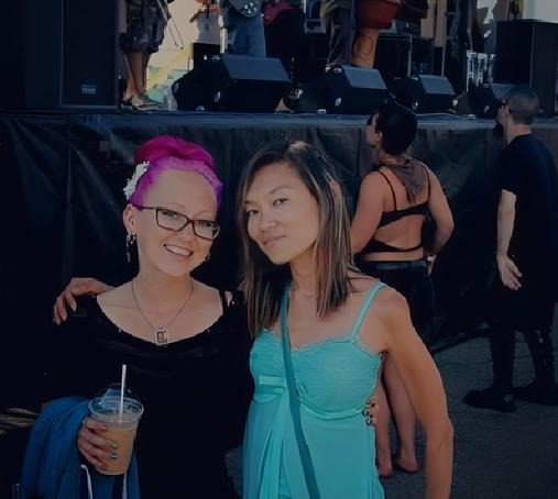 Two women, one with pink hair and glasses holding a drink, and the other(me) wearing a light blue chiffon tank top, smile at the camera, in front of an outdoor concert stage.