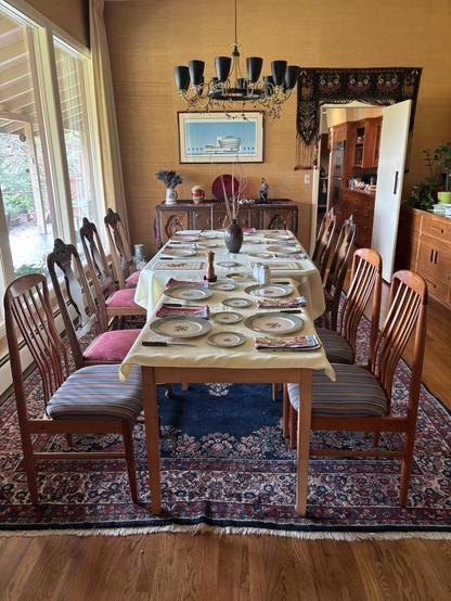 A dining room featuring two wooden tables joined together and set with plates and napkins. Wooden chairs surround the tables, and a decorative chandelier hangs above. The walls are painted in warm tones, and there is a rug beneath the table. Natural light streams through large windows