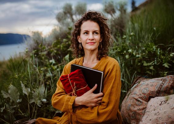 A woman with dark brown, curly hair sitting on a rock by a lake while wearing a yellow scarf hugs a book and smiles. Your favorite author would smile like that too if you helped make their publishing dreams a reality.
