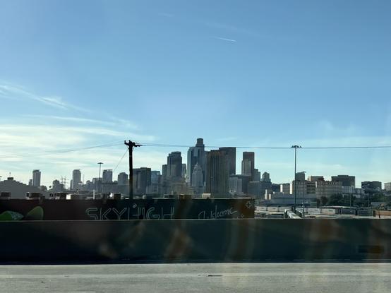 View of Downtown Los Angeles, captured in the late afternoon from the 5. City Hall is visible at the forefront of the skyline, only slightly. The sky itself is pale blue, thin wisps of cloud visible to the left of the image.