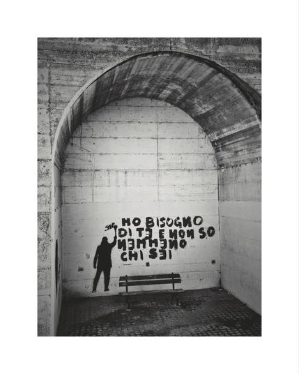 A vertical, black and white photograph of an arched concrete alcove. On the back wall, facing the viewer, there is a stencil or graffiti of a person's silhouette spray painting a graffiti. The text is in Italian. The legible part, painted in a black, thick, uneven font, reads: "HO BISOGNO DI TE E NON SO CHI SEI" which translates to "I NEED YOU AND I DON'T EVEN KNOW WHO YOU ARE". A simple metal bench with wooden slats sits below the graffiti, centered in the alcove. The floor is made of square pavers.