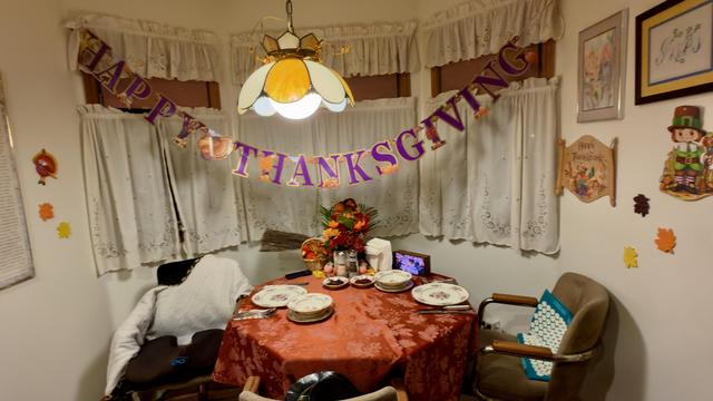 A Thanksgiving table for two set with a bronze tablecloth, fine china, two cranberry sauces, pumpkin and leaf decorations, and an autumnal flower arrangement. A Happy Thanksgiving banner in purple and gold hangs over the white drapes. Leaves are pasted to the wall.