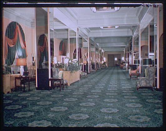The image depicts an elegant hotel lobby with a long, open layout and high ceilings. The flooring is covered in patterned carpeting that adds texture to the space. Along both sides of the hallway are rows of vintage-style chairs and couches adorned with plush cushions for comfort. Several tables hold lamps emitting warm light, creating a welcoming ambiance.

The room's walls are accented by tall, floor-to-ceiling mirrors framed with ornate molding, reflecting part of the interior and making the space appear larger. Curtains drape gracefully from ceiling to nearly the ground near each seating area, offering privacy while maintaining an open feel. The color palette is a mix of cool tones such as greens and blues on the walls and curtains, contrasted by warm hues like rusts in some chairs.

Overall, this hotel lobby exudes classic luxury with its attention to detail in furnishing and decor.