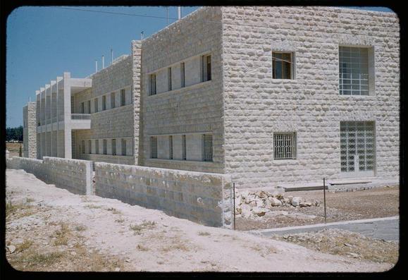 The image displays a large, multi-story building constructed from what appears to be concrete blocks. Its architecture is simple and functional with an emphasis on horizontal lines created by the window frames and eaves of the structure. The color palette consists mainly of muted tones such as off-white or beige for the walls, complemented by darker grays along some sections like ledges beneath windows.

The building's facade includes various types of openings: tall rectangular blocks likely serve as large glass panels while smaller barred windows provide security and additional ventilation in other areas. There is a noticeable absence of ornamental features which suggests utilitarian design possibly aimed at maximizing space or reflecting the era’s architectural trends favoring minimalism over ornamentation.

Adjacent to this building, there's an unpaved expanse with some scattered rocks and sparse vegetation indicative of arid conditions typical for many regions in Israel. A simple barrier made from large blocks separates this area from a paved path leading up to what might be another entrance or exit point associated with the hospital complex mentioned as "Jerusalem French Hospital on Mt. Scopus."

The overall setting appears dry, possibly due to environmental factors such as lack of rainfall or natural arid climate which could correspond to historical conditions in Jerusalem during mid-20th century considering the da [...]