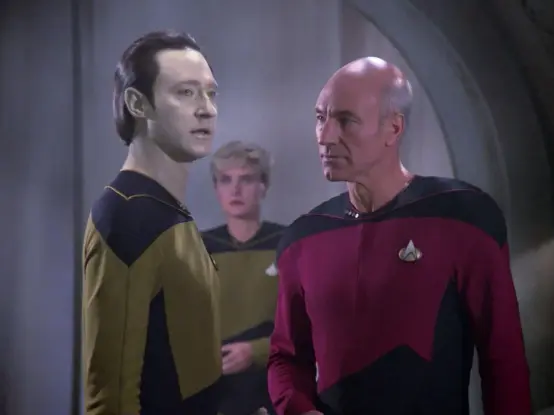 Brent Spiner appears as Data, Denise Crosby as Yar, and Patrick Stewart as Picard in Too Short a Season, And early episode of Star Trek: The Next Generation. The three Starfleet officers are standing in a dusty, rundown tunnel. Data is speaking, while the other two look on grimly.