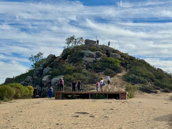 A view of Cerro San Luis peak. A couple of people are up at the top, silhouetted against the blue sky with wispy clouds. Below, there is a wooden deck with a group of people what were singing and dancing to music. Cerro San Luis, San Luis Obispo, California.