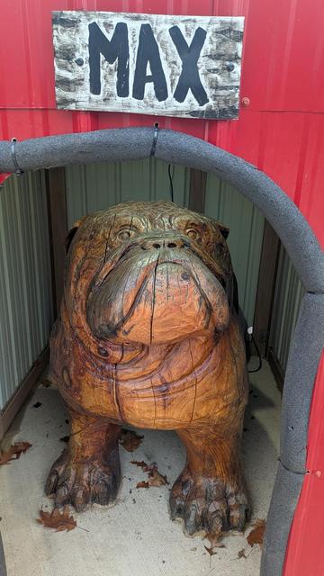 A photograph of a large, realistic wooden sculpture of an English bulldog sitting inside a custom red and black dog house. The dog house has a sign above the arched doorway that says "MAX" in white lettering. 