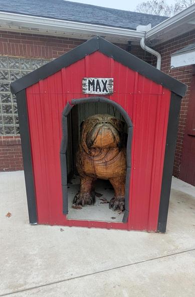 A photograph of a large, realistic wooden sculpture of an English bulldog sitting inside a custom red and black dog house. The dog house has a sign above the arched doorway that says "MAX" in white lettering.