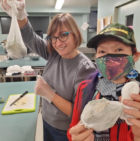 Two women in a commercial kitchen setting hold up small, filled, white mesh bags. The woman on the left, wearing eyeglasses and a gray sweater, smiles and raises a bag towards the camera while wearing clear gloves. Me, on the right, wearing a "AnarchoSocialist" hat and sequin mask, also holds a bag, wearing a Palestinian scarf and embroidered flower Asian earrings. A cutting board with a knife and chopped vegetables is on the counter in the foreground.