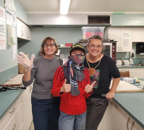 Three smiling women posing in a kitchen, with me, in the center wearing a sequined face mask, giving a thumbs up, with the other two smiling and one wearing a clear glove, kitchen cabinets and appliances in the background.