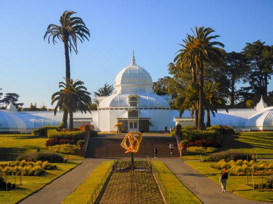 Symmetric composition showing an elaborate white Victorian greenhouse, flanked by palm trees. People were walking in the plaza and on the stairs in front of the greenhouse.