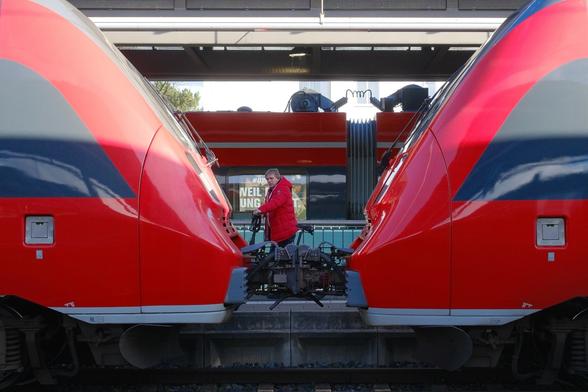 Ein roter Zug in Doppeltraktion steht an einem Bahnsteig. Der Blick geht zwischen den beiden Triebköpfen hindurch. Auf dem Bahnsteig dahinter schiebt ein Mann mit Jacke in passendem Rot ein Fahrrad. Er hat den Fotografen bemerkt und schaut mit verhaltenem Ausdruck in die Kamera. Im Hintergrund steht ein weiterer roter Zug. A red train in double traction stands at a platform. The view extends between the two power cars. On the platform behind it, a man wearing a matching red jacket pushes a bicycle. He has noticed the photographer and looks into the camera with a reserved expression. Another red train stands in the background.