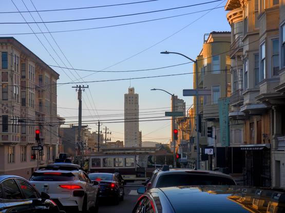 Cars were congested on a small street with cute Victorian row houses, waiting for a red light. A cable car was in the intersection. A white high-rise building is in the distance further down the street.