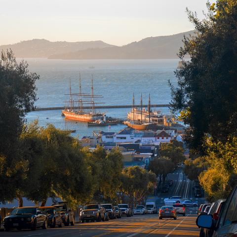 Looking down a steep road with cable car tracks, which leads to Fisherman's Wharf below where a cable car terminal is located. Two historical ships are docked at the wharf. Hills on the other side of the bay look hazy.