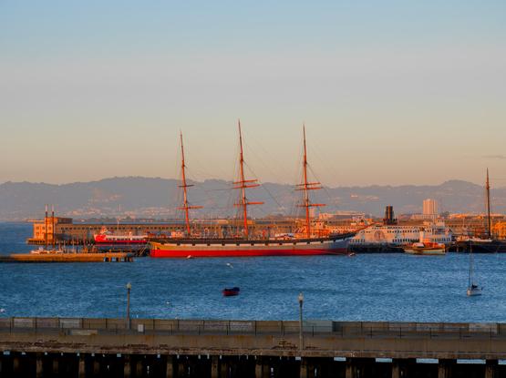 A historical ship with tall masts and some other smaller vessels are docked by Fisherman's Wharf, in the golden afternoon sun. Hazy hills on the other side of the bay can be seen.