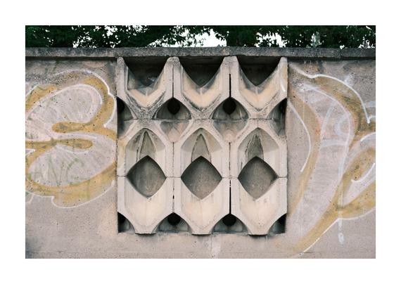 Betonwand mit einem eingelassenen, geometrisch geformten Sichtmuster aus wiederholten diamant- und bogenförmigen Elementen. Die Oberfläche ist verwittert und von hellgelbem und weißem Graffiti umgeben. Hinter den Öffnungen ist dunkler Hohlraum zu erkennen, darüber ragen Baumkronen in den Himmel.