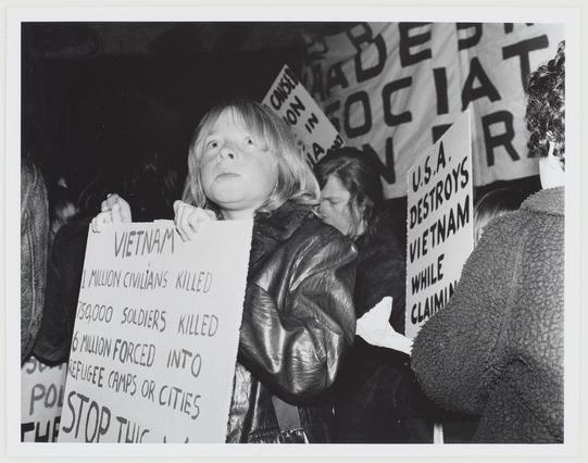 The photograph captures a moment from an anti-Vietnam War protest. A young girl with long hair is holding up a large sign with the heading "VIETNAM" prominently displayed at the top in bold, capitalized letters. Below it are several statistics: "3 million civilians killed," "560,000 soldiers killed," and other related figures such as "12 million forced into refugee camps or cities." The text on her sign is black against a white background, contrasting starkly with the girl's light skin tone.

In the background, various protest signs can be seen held by individuals participating in the demonstration. One prominent sign reads "U.S.A. DESTROYS VIETNAM WHILE CLAIMING," suggesting that there are more signs or messages just out of view. The crowd appears to include people with different expressions and postures, indicative of a peaceful assembly.

The image is presented as part of an article on anti-Vietnam war demonstrations, which can be found at the provided URL: Anti Vietnam war demonstration (https://images.loener.nl/AnsWestra/full/6690/6690f4a63c8dcd5ca3a70888.jpg). The context and historical significance of this image are linked to the broader social, political, and cultural movements against American involvement in Vietnam during that era.